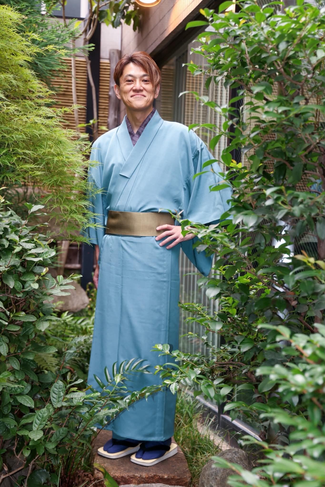 庭の緑に囲まれ、水色の着物と金茶色の帯をまとった藤本大士さんの全身姿