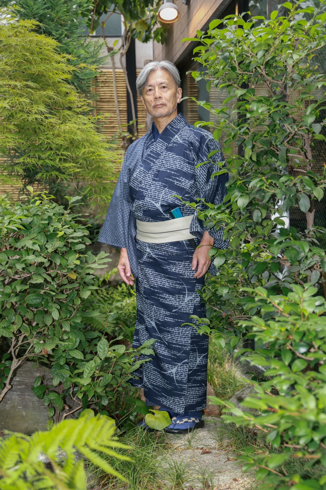 庭の緑に囲まれ、紺色の着物を着た丸地典利さんの全身姿